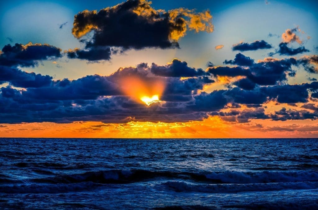 Sun setting over dark blue ocean waves, with orange light breaking through a bank of dramatic clouds.
