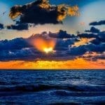 Sun setting over dark blue ocean waves, with orange light breaking through a bank of dramatic clouds.