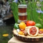 Three stacked jars of tomato jam sit beside fresh tomatoes, a lime, a pepper and crackers topped with jam on a wooden board.