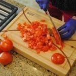 Hands in blue gloves dice fresh red tomatoes on a wooden cutting board beside whole tomatoes and a gas stove.