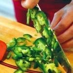 A person slices green jalapeño peppers on a wooden cutting board with a chef's knife.