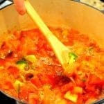 A hand stirs chunky diced tomatoes, peppers and onions simmering in a blue enameled Dutch oven with a wooden spoon.
