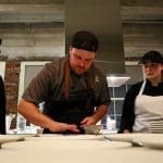 Four cooks in black and aprons stand at a counter of white plates while a chef plates food in a brick-walled kitchen.