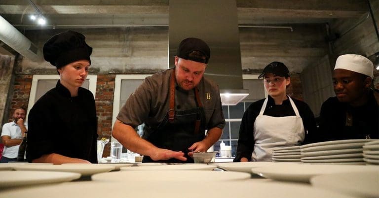 Four cooks in black and aprons stand at a counter of white plates while a chef plates food in a brick-walled kitchen.