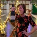 A smiling woman in a black and red floral dress holds up a bottle of wine at an indoor evening reception.