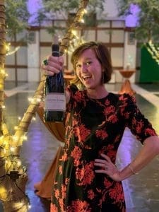 A smiling woman in a black and red floral dress holds up a bottle of wine at an indoor evening reception.