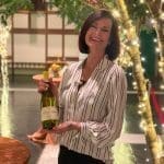 Smiling woman with short auburn hair holds a bottle of Sonoma-Cutrer chardonnay beneath twinkling lights and trees.