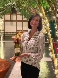 Smiling woman with short auburn hair holds a bottle of Sonoma-Cutrer chardonnay beneath twinkling lights and trees.