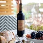 A bottle of Cabernet Franc wine beside an empty glass, grapes, olives, and bread on a wooden outdoor table.