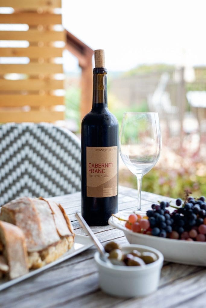 A bottle of Cabernet Franc wine beside an empty glass, grapes, olives, and bread on a wooden outdoor table.