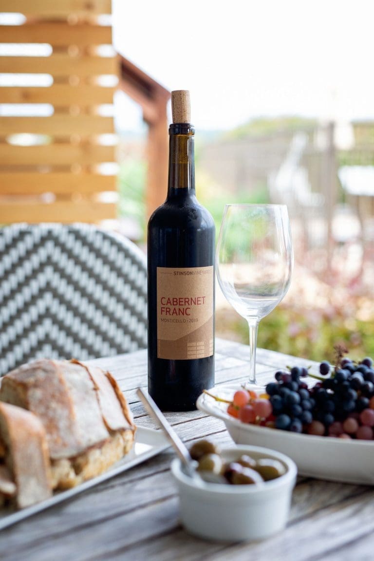 A bottle of Cabernet Franc wine beside an empty glass, grapes, olives, and bread on a wooden outdoor table.
