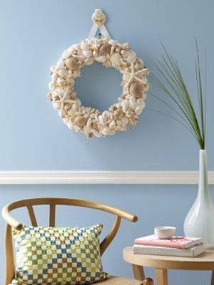 A seashell wreath with starfish hangs on a light blue wall above a wooden chair and a side table with a grass vase.