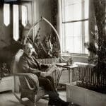 A vintage black-and-white photo of a mustached man reading a newspaper in a wicker rocking chair by a sunlit window.
