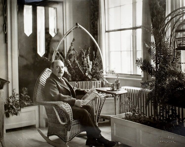 A vintage black-and-white photo of a mustached man reading a newspaper in a wicker rocking chair by a sunlit window.
