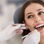 A smiling woman in a dental chair while a gloved dentist examines her teeth with a mirror and probe.