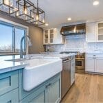 A bright kitchen with white shaker cabinets, a teal island with a farmhouse sink, and glass pendant lights.