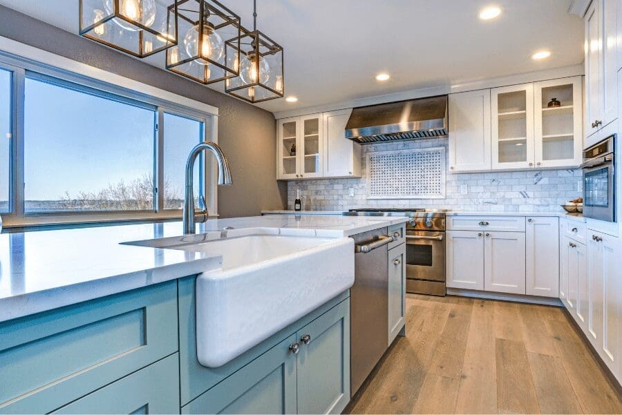 A bright kitchen with white shaker cabinets, a teal island with a farmhouse sink, and glass pendant lights.