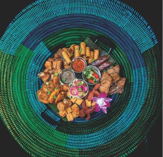 Overhead view of a Filipino appetizer platter with spring rolls, fried pork, sausages, dipping sauces and an orchid on a colorful woven mat.