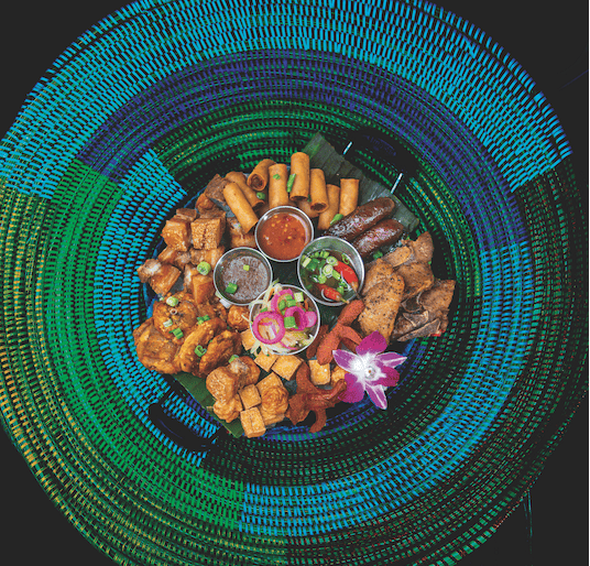 Overhead view of a Filipino appetizer platter with spring rolls, fried pork, sausages, dipping sauces and an orchid on a colorful woven mat.