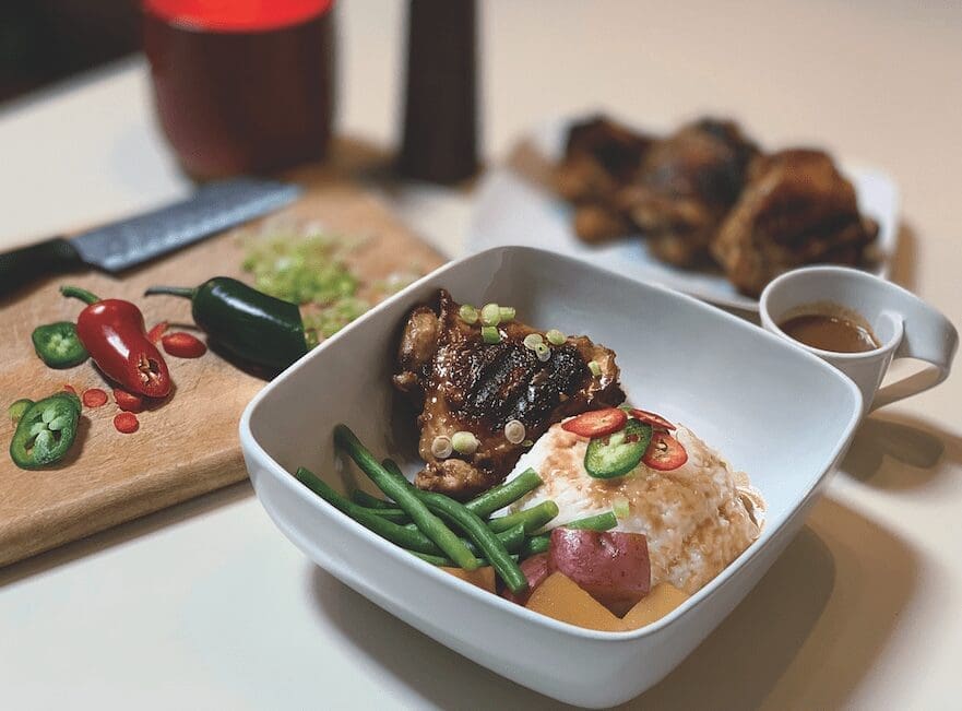 A white square bowl holds glazed chicken thighs, rice, green beans and potatoes, with sliced chilies and a small sauce cup.