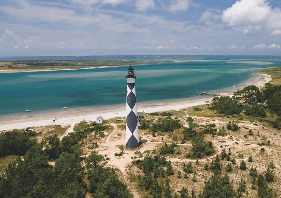 A black-and-white diamond-patterned lighthouse stands on sandy dunes beside turquoise water and a sandy beach, viewed from above.