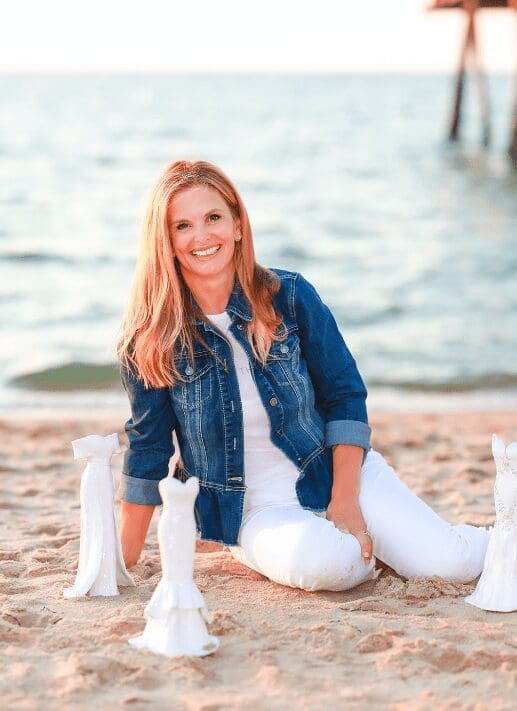 A smiling woman with long blonde hair in a denim jacket and white jeans sits on a sandy beach beside white ceramic vases.