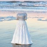 A white gown with a sparkly beaded bodice stands on wet sand at the water's edge at sunset, with no visible person.