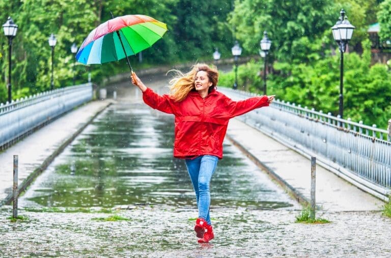 A woman in a red raincoat and red boots dances on a wet bridge under a rainbow umbrella.