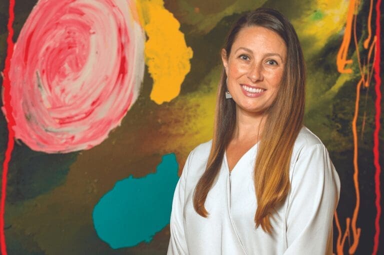 A smiling woman with long light brown hair in a white top, standing before a colorful abstract painting with pink, yellow and teal shapes.