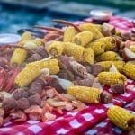 A spread of seafood boil on a red-and-white checkered tablecloth with shrimp, corn on the cob, sausage, potatoes and crab legs.