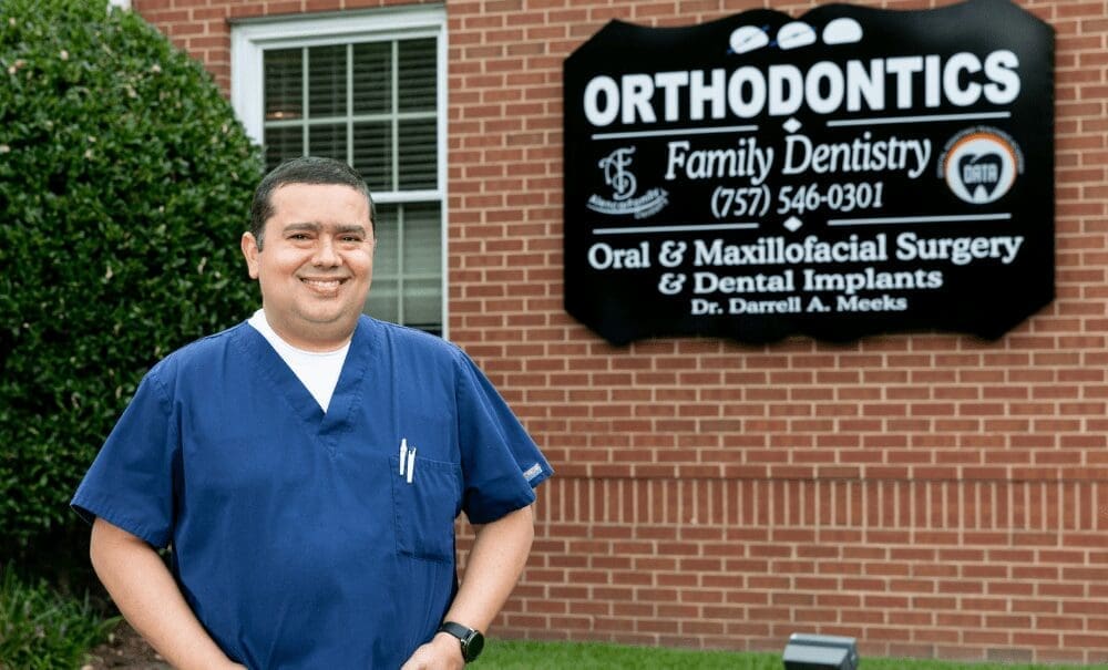 A smiling man in blue scrubs stands before a brick wall beside a black sign reading "Orthodontics, Family Dentistry, Oral & Maxillofacial Surgery.
