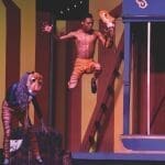 A stage performance with actors in tiger and wolf-like masks, one leaping shirtless in orange shorts beside a striped circus-style set.