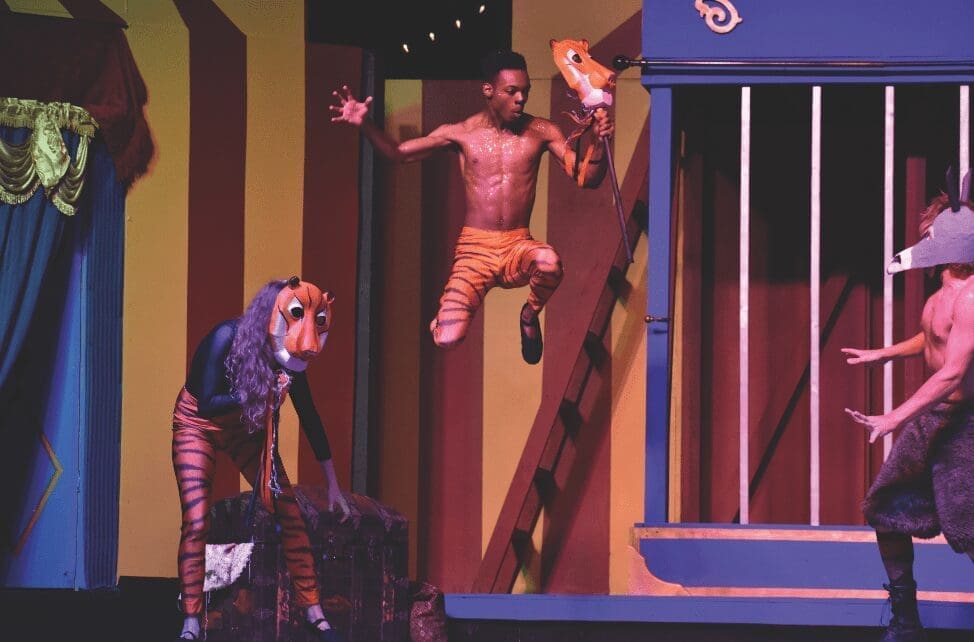 A stage performance with actors in tiger and wolf-like masks, one leaping shirtless in orange shorts beside a striped circus-style set.