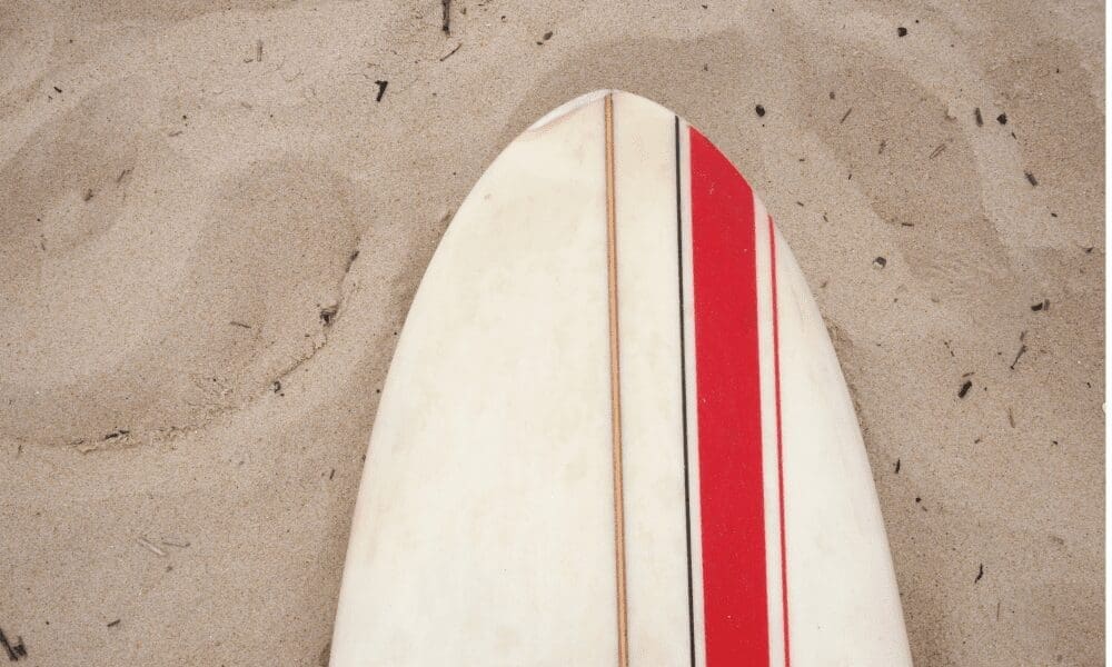 A white surfboard with red and thin pinstripes lies nose-first in the sand.