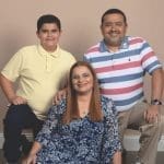 Three smiling family members posed against a beige backdrop: a teen boy in yellow, a woman in a blue floral dress, and a man in a striped polo.