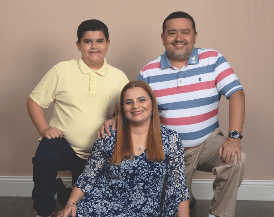 Three smiling family members posed against a beige backdrop: a teen boy in yellow, a woman in a blue floral dress, and a man in a striped polo.