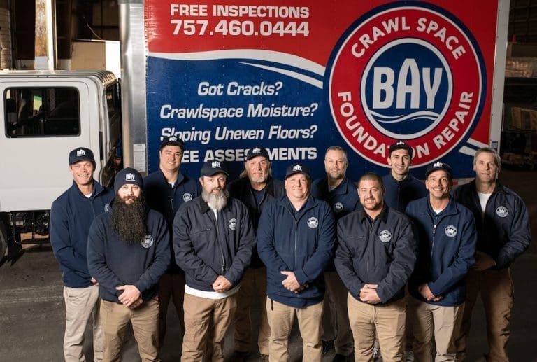 A group of ten men in navy jackets stands before a BAY Crawl Space Foundation Repair truck with a "Free Inspections" sign.