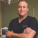 Smiling man in dark scrubs with arms crossed in an exam room, scrubs reading Mancoll Cosmetic & Plastic Surgery.