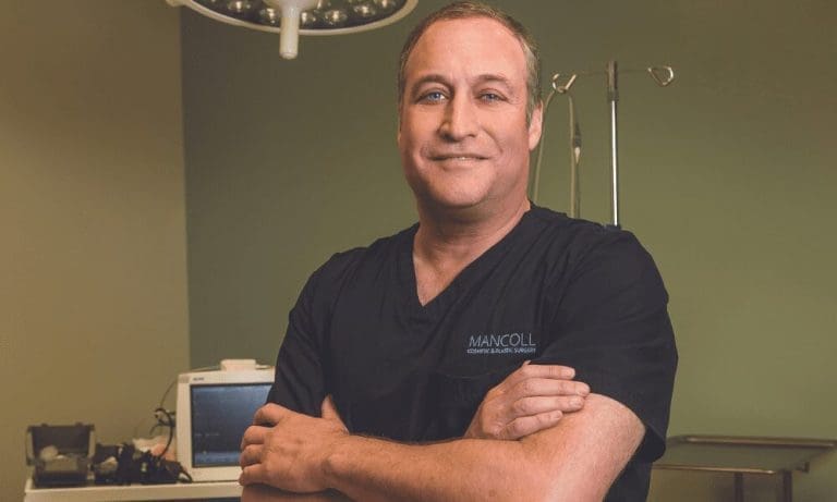 Smiling man in dark scrubs with arms crossed in an exam room, scrubs reading Mancoll Cosmetic & Plastic Surgery.