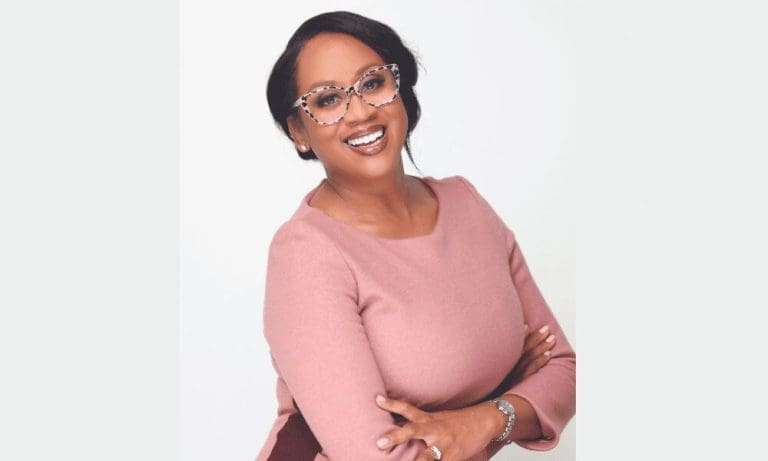 A smiling woman in cat-eye glasses and a pink long-sleeve top, arms crossed, against a white background.