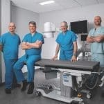 Four men in blue and teal scrubs pose beside an operating table and medical imaging equipment in a bright room.