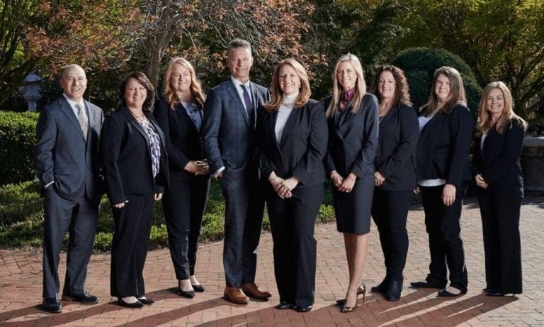 Nine professionals in dark business attire pose together on a brick walkway outside a building with greenery.