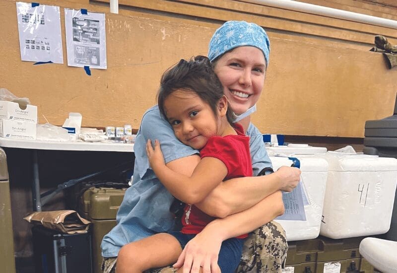 A smiling woman in blue scrubs and a surgical cap hugs a young girl in a red shirt in a storage room.
