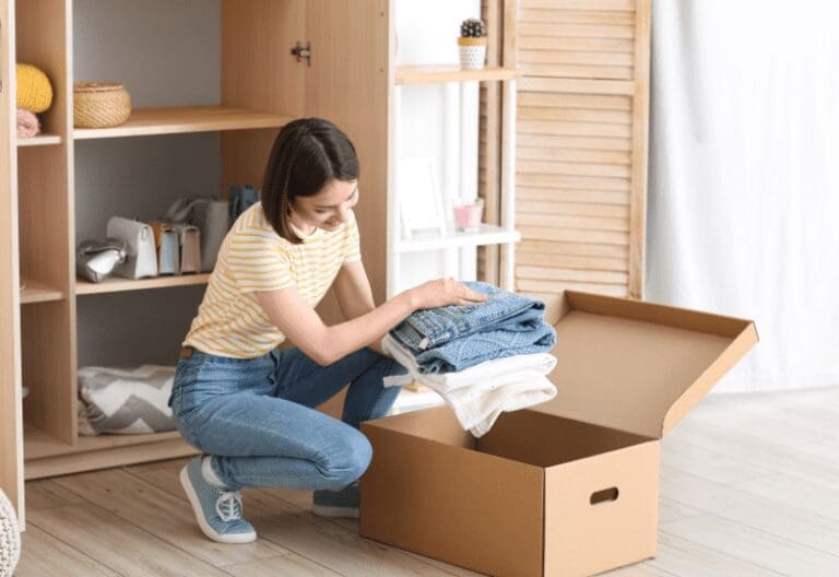 A woman in a striped yellow top kneels and packs folded jeans and white clothes into an open cardboard box beside a wooden closet.