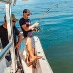 Two men on a boat's bow fish with a bent rod over calm blue water, while a net waits near the rail.
