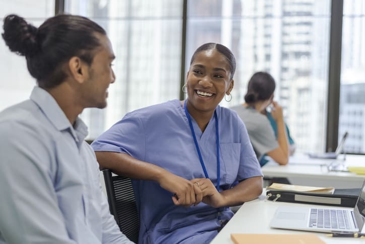 A black female student smiles at her classmate. Sitting at a desk in a modern classroom setting. Laptop on desk with note boo