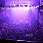 A packed crowd fills a dimly lit venue under purple stage lights, watching a band perform from the balconies above.