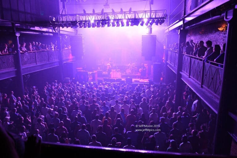 A packed crowd fills a dimly lit venue under purple stage lights, watching a band perform from the balconies above.