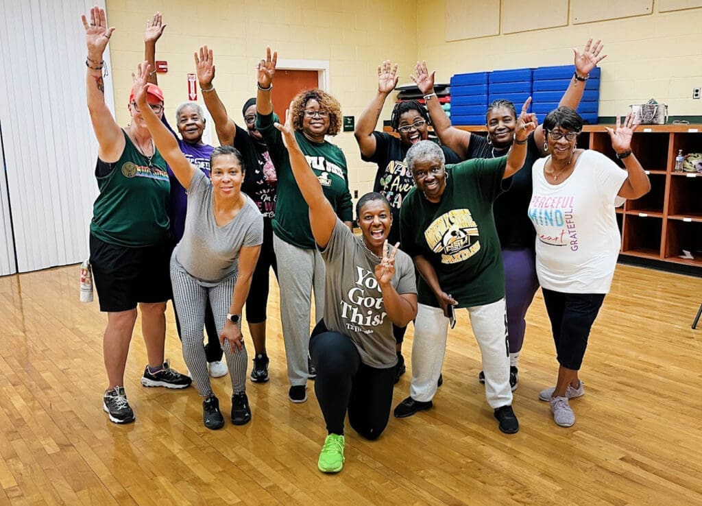 A group of smiling women in green and gray shirts raise their arms in a gym with wood floors.