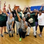 A group of smiling women in green and gray shirts raise their arms in a gym with wood floors.